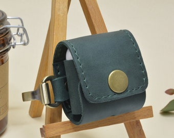 May include: A dark green leather case for wireless earbuds with a gold metal clasp and a gold button. The case is sitting on a small wooden easel.