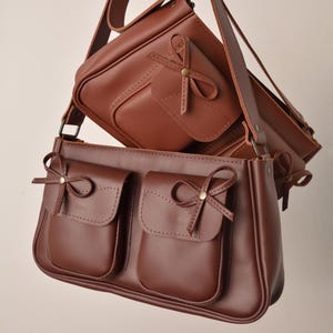 May include: Two brown leather crossbody bags. The larger bag has two front pockets with bow accents. The smaller bag is positioned above, also with a bow accent. Both bags have adjustable straps and a classic, elegant design. The bags are made of a smooth, rich brown leather.