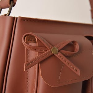 Pu&ograve; includere: Primo piano di una borsa a tracolla in pelle marrone con un dettaglio a fiocco sulla tasca anteriore. La borsa ha una chiusura con patta e una tracolla. Le cuciture sono visibili e l'hardware &egrave; dorato.