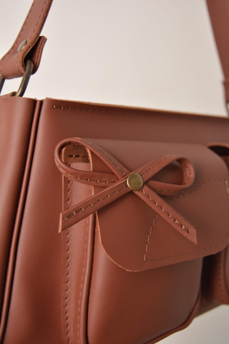 Pu&ograve; includere: Primo piano di una borsa a tracolla in pelle marrone con un dettaglio a fiocco sulla tasca anteriore. La borsa ha una chiusura con patta e una tracolla. Le cuciture sono visibili e l'hardware &egrave; dorato.