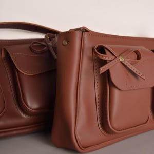 Pu&ograve; includere: Due borse a mano in pelle marrone con dettagli a fiocco. Le borse hanno una forma rettangolare con una tasca frontale con patta. Sono realizzate in pelle liscia e opaca, con un aspetto classico e vintage.