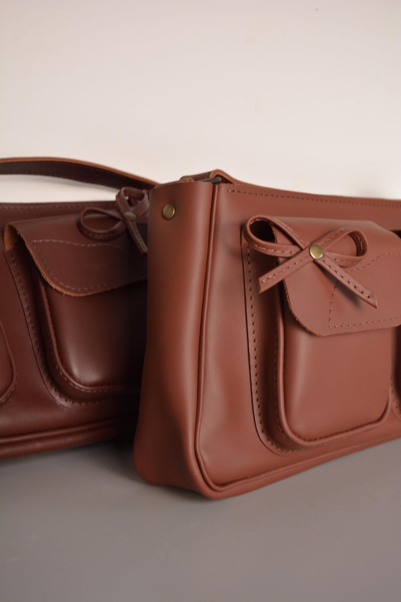 Pu&ograve; includere: Due borse a mano in pelle marrone con dettagli a fiocco. Le borse hanno una forma rettangolare con una tasca frontale con patta. Sono realizzate in pelle liscia e opaca, con un aspetto classico e vintage.