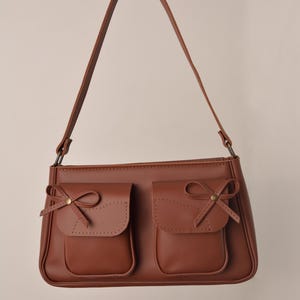 Pu&ograve; includere: Una borsa marrone con due tasche frontali, ognuna ornata da un piccolo fiocco. La borsa ha una tracolla lunga e sottile e una forma rettangolare. Il colore &egrave; un marrone medio caldo.