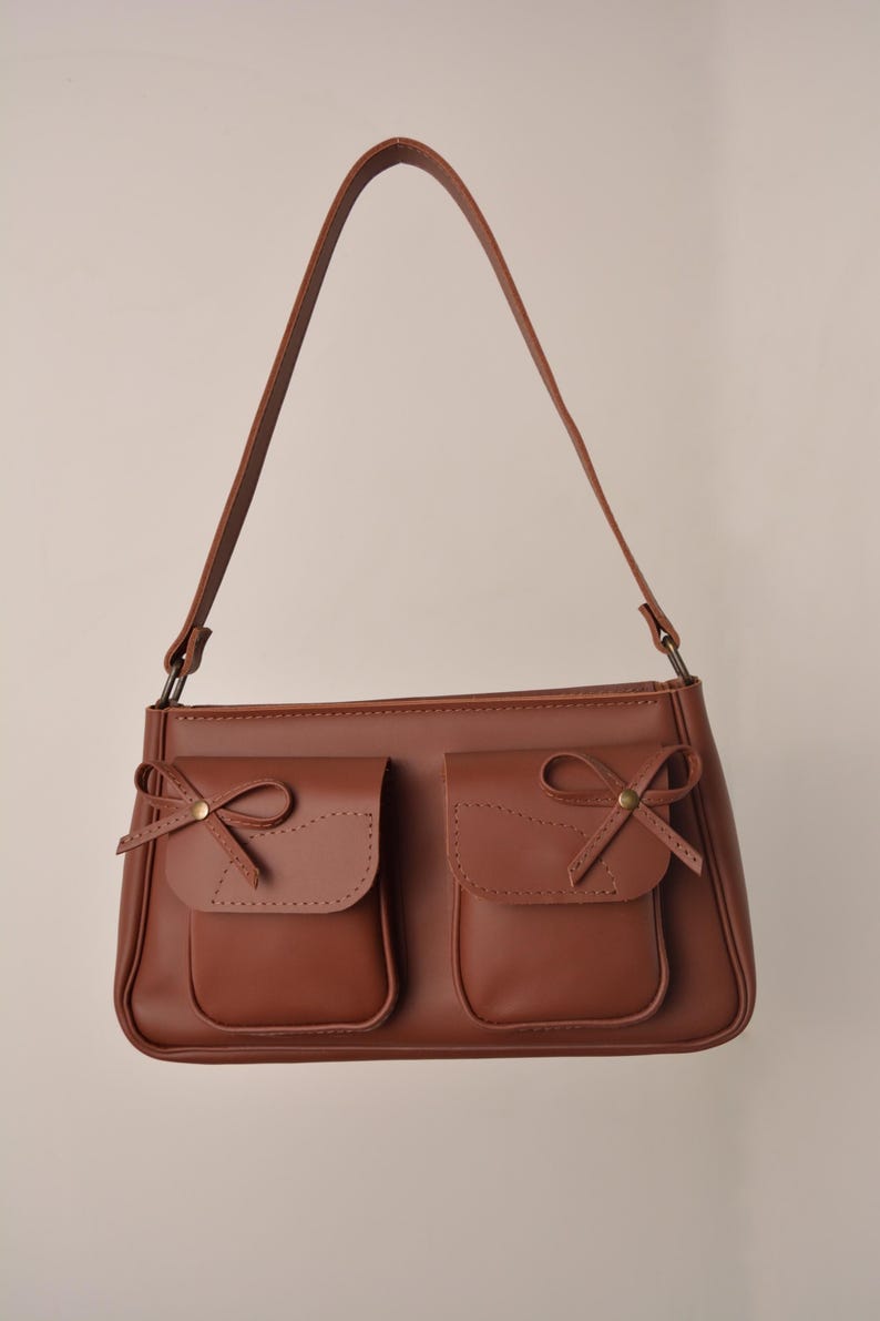 Pu&ograve; includere: Una borsa marrone con due tasche frontali, ognuna ornata da un piccolo fiocco. La borsa ha una tracolla lunga e sottile e una forma rettangolare. Il colore &egrave; un marrone medio caldo.