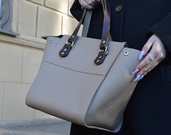 May include: A beige leather tote bag with brown leather handles and silver hardware. The bag has a zipper closure and a flap with a magnetic closure.