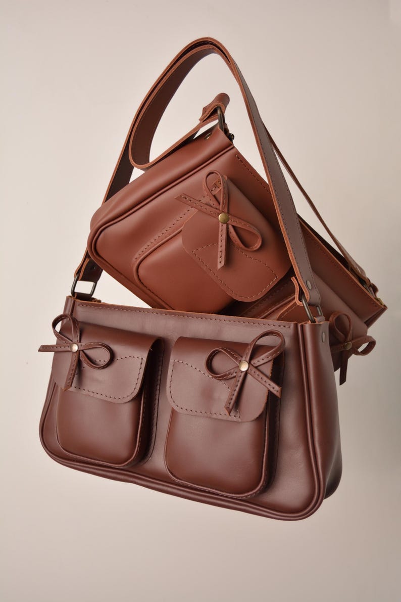 May include: Two brown leather crossbody bags. The larger bag has two front pockets with bow accents. The smaller bag is positioned above, also with a bow accent. Both bags have adjustable straps and a classic, elegant design. The bags are made of a smooth, rich brown leather.