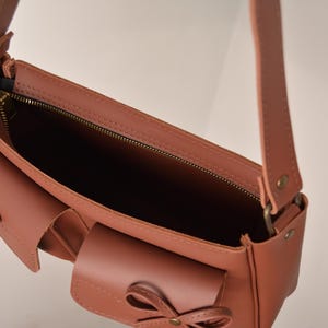 Pu&ograve; includere: Borsa a tracolla in pelle color rosa antico con una lunga tracolla e due tasche frontali. La borsa ha una chiusura a zip e dettagli a fiocco decorativi sulle tasche. L'interno della borsa &egrave; visibile, mostrando una fodera scura.