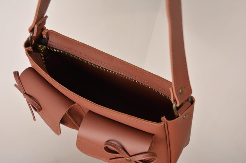 Pu&ograve; includere: Borsa a tracolla in pelle color rosa antico con una lunga tracolla e due tasche frontali. La borsa ha una chiusura a zip e dettagli a fiocco decorativi sulle tasche. L'interno della borsa &egrave; visibile, mostrando una fodera scura.