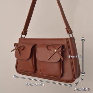 Pu&ograve; includere: Borsa a tracolla marrone con due tasche frontali, ciascuna ornata da un fiocco. La borsa misura 31 cm di larghezza, 18 cm di altezza e 7 cm di profondit&agrave;. Ha una lunga tracolla.