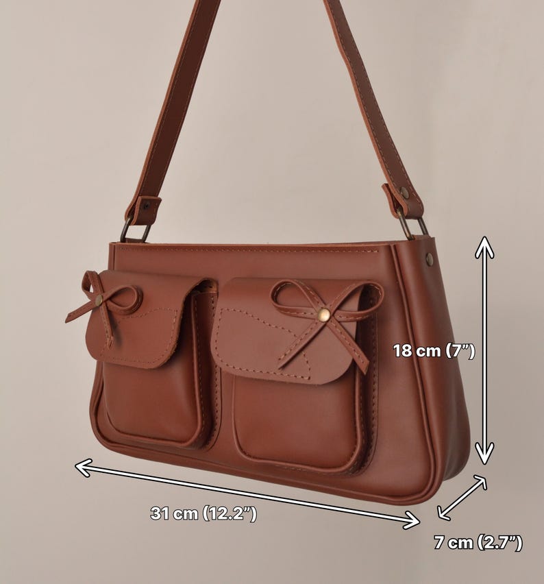 Pu&ograve; includere: Borsa a tracolla marrone con due tasche frontali, ciascuna ornata da un fiocco. La borsa misura 31 cm di larghezza, 18 cm di altezza e 7 cm di profondit&agrave;. Ha una lunga tracolla.