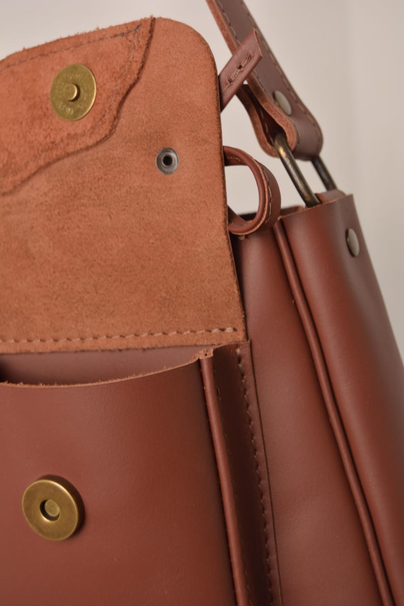 Pu&ograve; includere: Borsa a tracolla in pelle marrone con chiusura a patta e chiusura magnetica. La borsa ha un design strutturato con cuciture a vista e finiture metalliche. Il cinturino &egrave; fissato con anelli metallici.