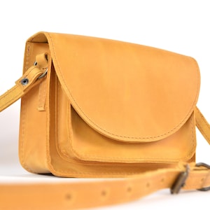May include: A mustard-yellow leather crossbody bag with a flap closure. The bag has a long, adjustable strap and a small front pocket. The bag is made of smooth leather and has a simple, classic design.
