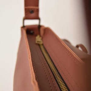 Pu&ograve; includere: Primo piano di una borsa in pelle marrone con cerniera in ottone. La borsa ha una tracolla ed &egrave; realizzata in pelle liscia e tinta unita. La cerniera &egrave; aperta, rivelando l'interno della borsa.