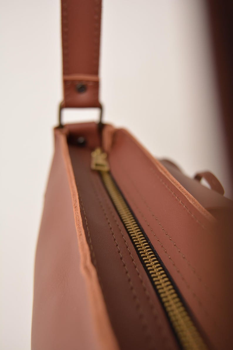 Pu&ograve; includere: Primo piano di una borsa in pelle marrone con cerniera in ottone. La borsa ha una tracolla ed &egrave; realizzata in pelle liscia e tinta unita. La cerniera &egrave; aperta, rivelando l'interno della borsa.