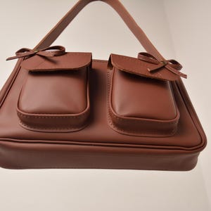 Pu&ograve; includere: Una borsa marrone con due tasche frontali, ciascuna ornata da un piccolo fiocco. La borsa ha una forma rettangolare con un manico ed &egrave; realizzata in un materiale liscio simile alla pelle. Il design &egrave; classico ed elegante.