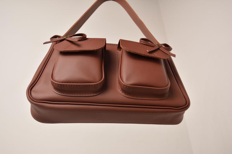 Pu&ograve; includere: Una borsa marrone con due tasche frontali, ciascuna ornata da un piccolo fiocco. La borsa ha una forma rettangolare con un manico ed &egrave; realizzata in un materiale liscio simile alla pelle. Il design &egrave; classico ed elegante.