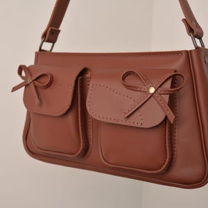 Pu&ograve; includere: Borsa a tracolla in pelle marrone con due tasche frontali, ciascuna ornata da un fiocco decorativo. La borsa ha una lunga tracolla e una forma rettangolare. Le cuciture sono visibili.