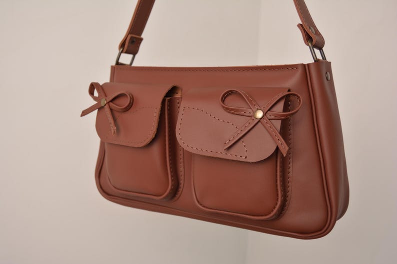 Pu&ograve; includere: Borsa a tracolla in pelle marrone con due tasche frontali, ciascuna ornata da un fiocco decorativo. La borsa ha una lunga tracolla e una forma rettangolare. Le cuciture sono visibili.