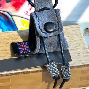 May include: A grey leather key chain with a black button and two silver metal beads with a Celtic knot design. The key chain has a small black tag with a Union Jack flag.