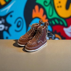 May include: Miniature brown leather boots with white stitching and laces. The boots have a white sole and are attached to a keychain. The background features colorful graffiti art.