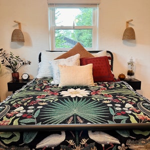 May include: A black bed with a white and green floral patterned throw blanket. The bed has a black metal frame and a white headboard. There are several pillows on the bed, including a white pillow with a brown and white patterned pillow on top. There are two wicker pendant lights on the wall behind the bed.