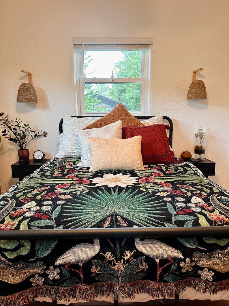 May include: A black bed with a white and green floral patterned throw blanket. The bed has a black metal frame and a white headboard. There are several pillows on the bed, including a white pillow with a brown and white patterned pillow on top. There are two wicker pendant lights on the wall behind the bed.