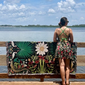 May include: A woman stands on a wooden dock, holding a woven tapestry with a floral and animal design. The tapestry features a variety of plants, flowers, and birds, including a white bird with a long beak. The tapestry is black with green, red, and white accents. The woman is wearing a floral dress and sunglasses. The background is a blue sky with white clouds and a body of water.