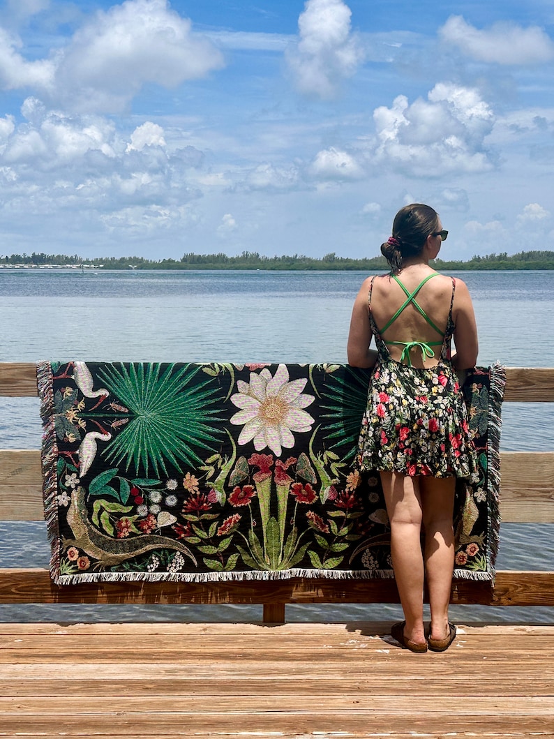 May include: A woman stands on a wooden dock, holding a woven tapestry with a floral and animal design. The tapestry features a variety of plants, flowers, and birds, including a white bird with a long beak. The tapestry is black with green, red, and white accents. The woman is wearing a floral dress and sunglasses. The background is a blue sky with white clouds and a body of water.