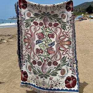 May include: A rectangular woven throw blanket featuring a symmetrical design with two large octopuses, pomegranates, fish, butterflies, and deep red floral motifs on a cream background, finished with a dark blue scalloped border and fringe.