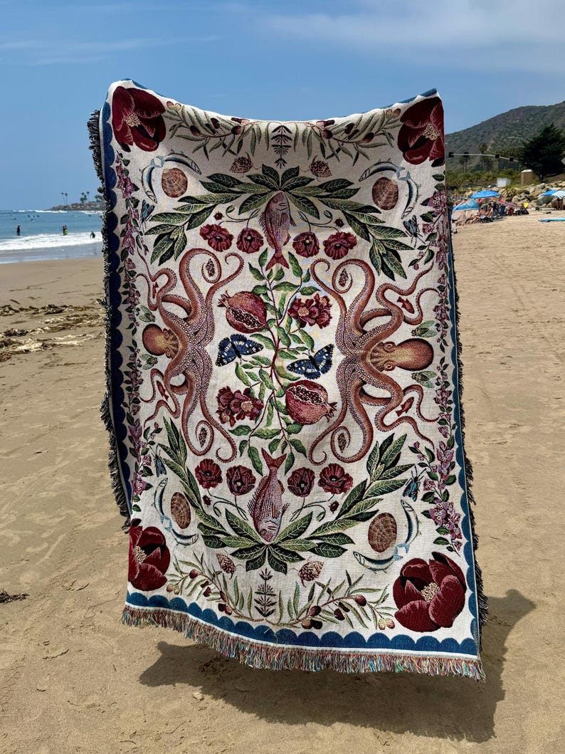May include: A rectangular woven throw blanket featuring a symmetrical design with two large octopuses, pomegranates, fish, butterflies, and deep red floral motifs on a cream background, finished with a dark blue scalloped border and fringe.