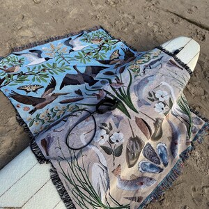 May include: A woven throw blanket featuring a split-colour design with a light blue section depicting birds, butterflies, and foliage, and a beige section showing marine life including fish, shells, a heron, and a horseshoe crab, with fringe edges.