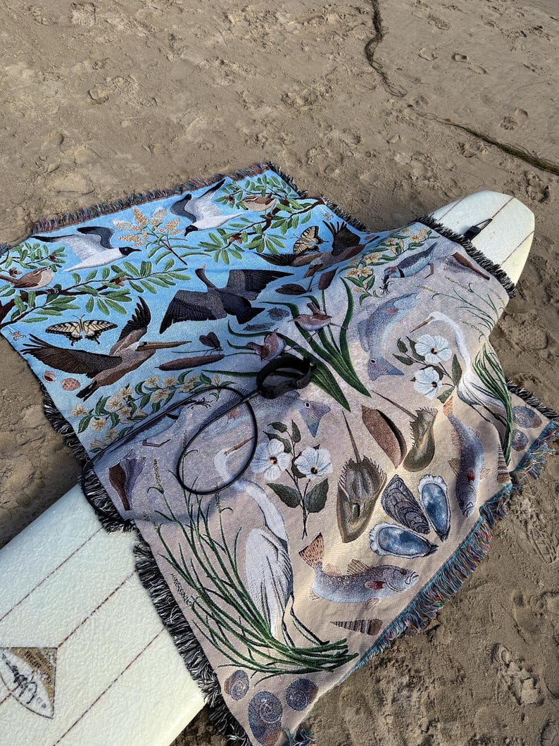 May include: A woven throw blanket featuring a split-colour design with a light blue section depicting birds, butterflies, and foliage, and a beige section showing marine life including fish, shells, a heron, and a horseshoe crab, with fringe edges.