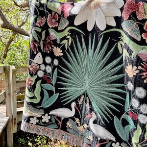 May include: A black woven tapestry throw blanket with a colorful floral and bird design. The design features white birds, green palm leaves, and pink and white flowers.