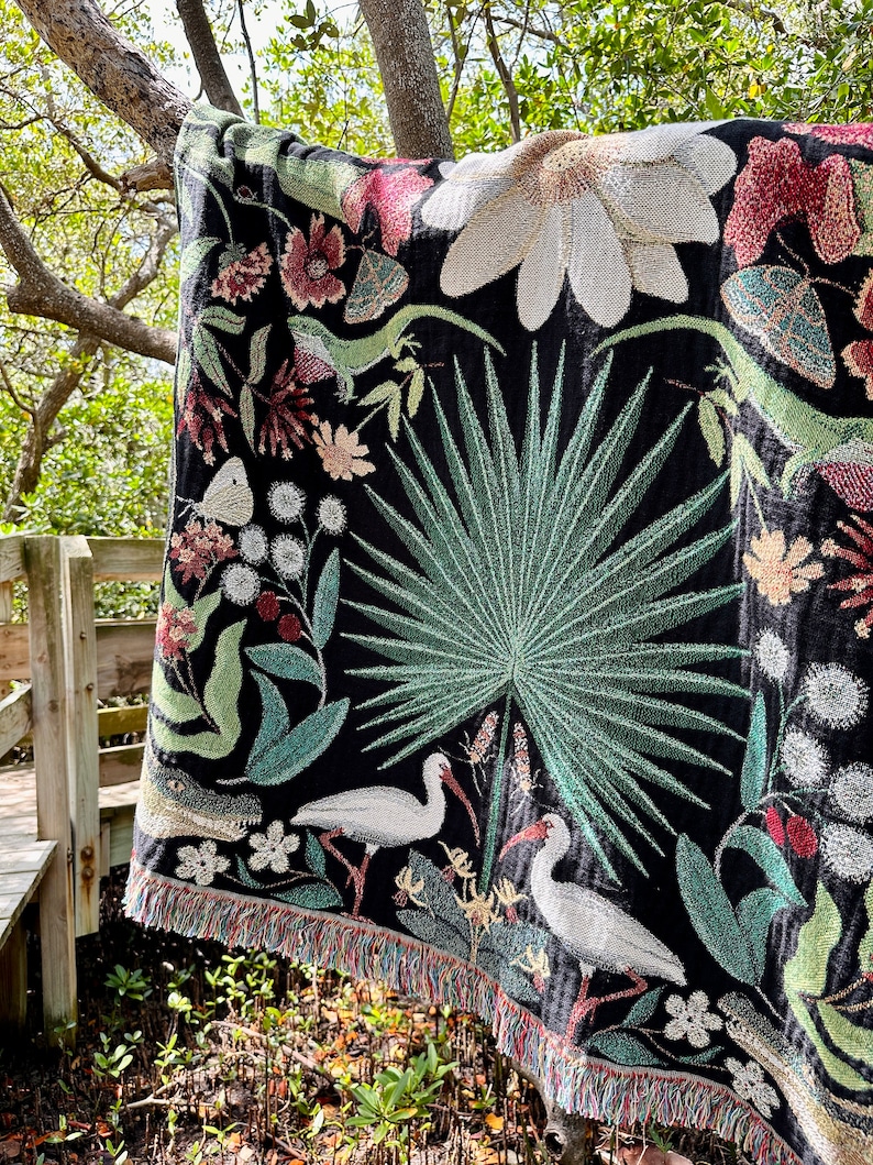 May include: A black woven tapestry throw blanket with a colorful floral and bird design. The design features white birds, green palm leaves, and pink and white flowers.