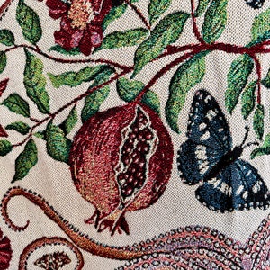 May include: A woven textile featuring a cream-colored background with a detailed pattern of red pomegranates, green leaves, dark red flowers, and blue butterflies. The design also includes a portion of an octopus tentacle with visible suction cups.