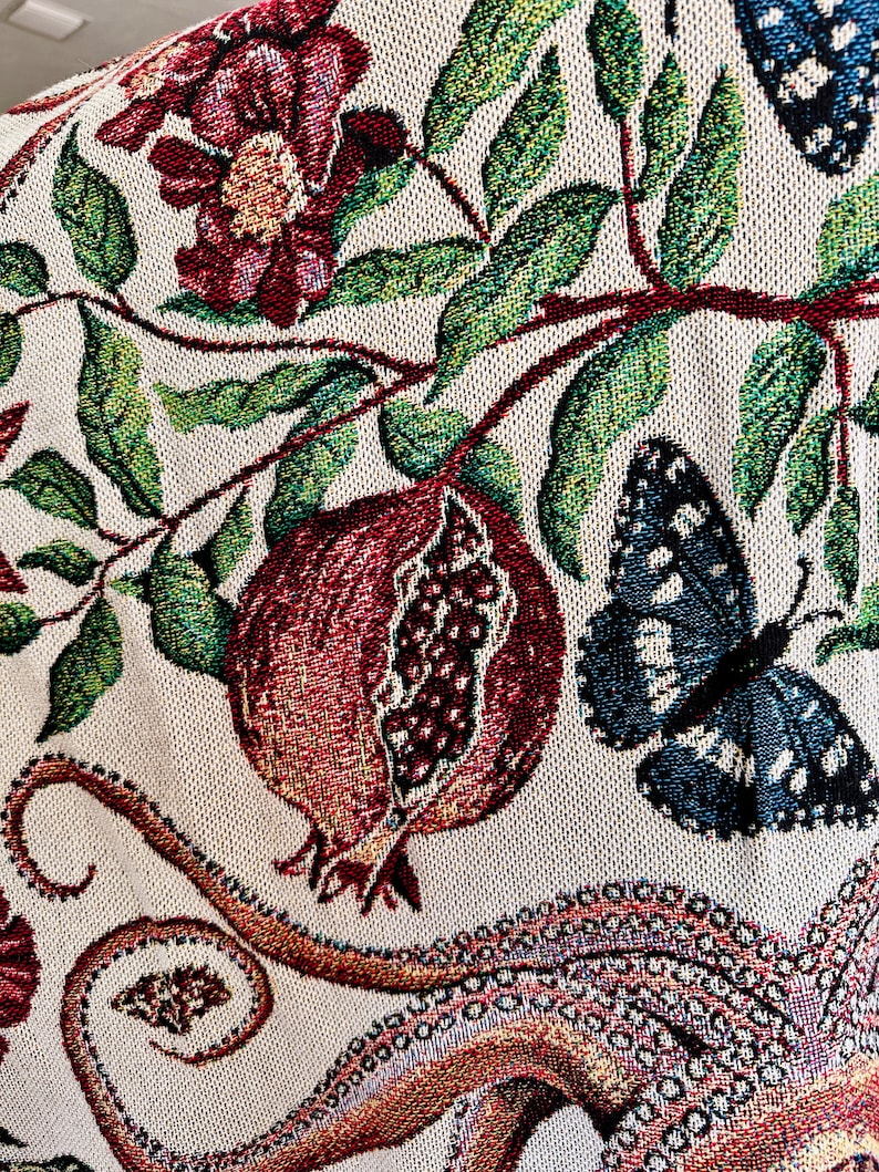 May include: A woven textile featuring a cream-colored background with a detailed pattern of red pomegranates, green leaves, dark red flowers, and blue butterflies. The design also includes a portion of an octopus tentacle with visible suction cups.