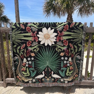 May include: A black woven throw blanket with a floral and animal design. The design features white birds, green palm leaves, and a white flower. The blanket has a fringed edge.