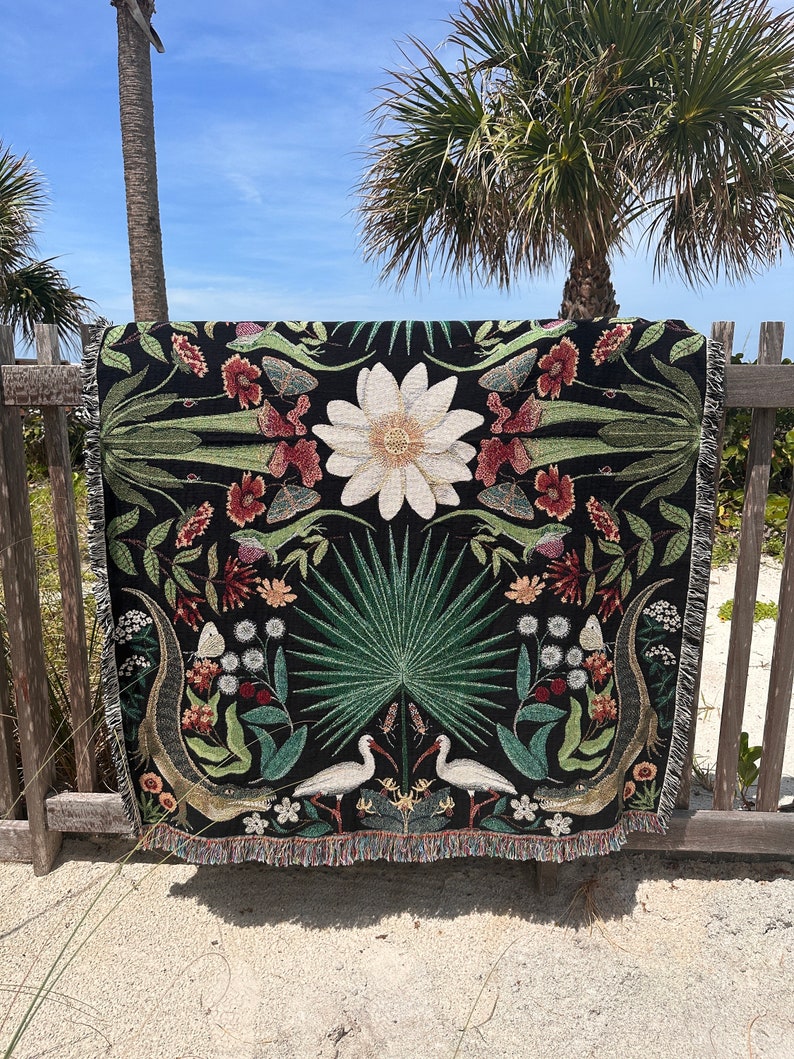 May include: A black woven throw blanket with a floral and animal design. The design features white birds, green palm leaves, and a white flower. The blanket has a fringed edge.