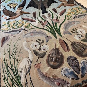 May include: A woven throw blanket featuring a detailed nature scene with a light blue sky and tan ground. The design includes a white egret, various fish, horseshoe crabs, oysters, pelicans, butterflies, dragonflies, and green marsh grasses.