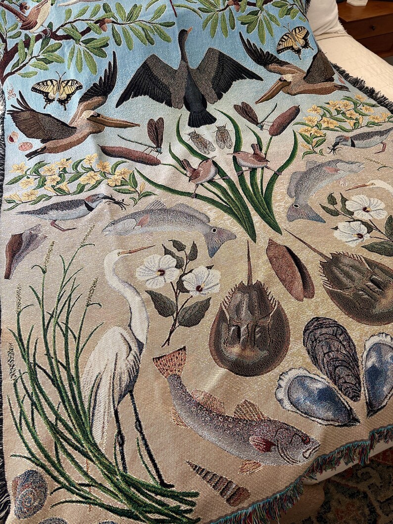 May include: A woven throw blanket featuring a detailed nature scene with a light blue sky and tan ground. The design includes a white egret, various fish, horseshoe crabs, oysters, pelicans, butterflies, dragonflies, and green marsh grasses.