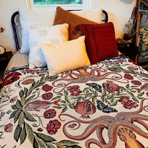 May include: A woven throw blanket featuring a detailed botanical and marine pattern with an octopus, fish, pomegranates, and butterflies in shades of red, green, and blue on a cream background, layered over a bed with several decorative pillows.
