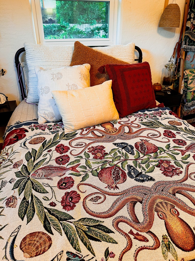 May include: A woven throw blanket featuring a detailed botanical and marine pattern with an octopus, fish, pomegranates, and butterflies in shades of red, green, and blue on a cream background, layered over a bed with several decorative pillows.