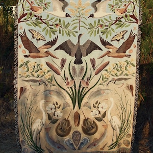 May include: A woven throw blanket featuring a symmetrical nature-themed design with birds, fish, butterflies, and aquatic life. The illustration includes pelicans, herons, horseshoe crabs, and various flora in shades of blue, brown, and cream.