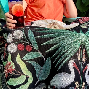 May include: A black and white woven throw blanket with a floral and tropical design. The blanket features white birds, green palm leaves, and red berries. The blanket is draped over a teal chair.
