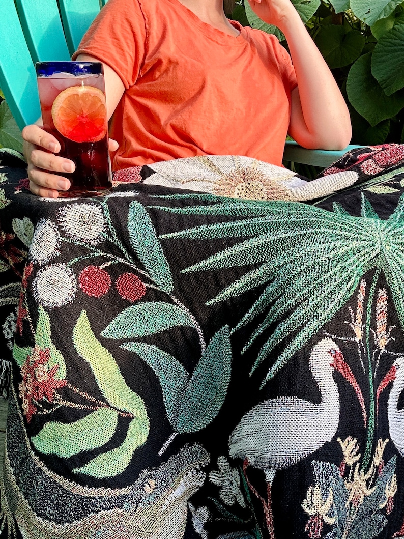 May include: A black and white woven throw blanket with a floral and tropical design. The blanket features white birds, green palm leaves, and red berries. The blanket is draped over a teal chair.
