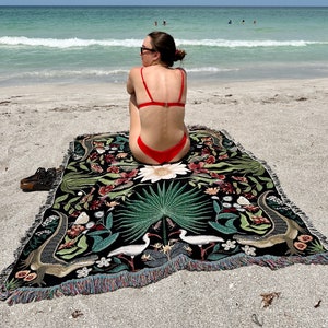 May include: A woman wearing a red bikini sits on a black and green tapestry with a floral and animal design. The tapestry has a fringe border. The woman is sitting on a sandy beach with the ocean in the background.