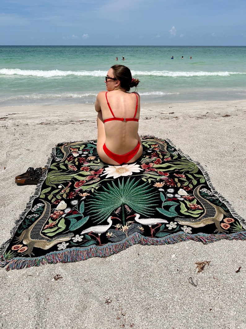May include: A woman wearing a red bikini sits on a black and green tapestry with a floral and animal design. The tapestry has a fringe border. The woman is sitting on a sandy beach with the ocean in the background.