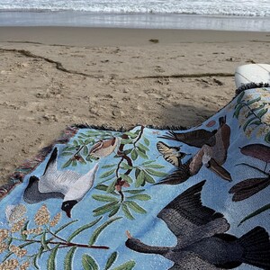 May include: A light blue woven throw blanket featuring a detailed pattern of various birds in flight, including a dark cormorant and a white gull, interspersed with green leafy branches, small brown birds, and a yellow butterfly with black markings.
