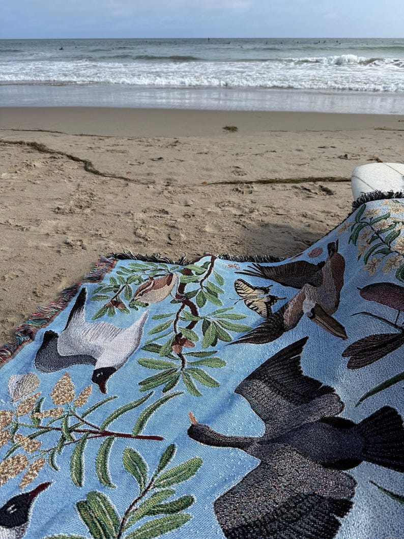 May include: A light blue woven throw blanket featuring a detailed pattern of various birds in flight, including a dark cormorant and a white gull, interspersed with green leafy branches, small brown birds, and a yellow butterfly with black markings.