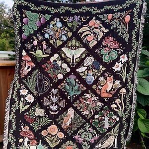 May include: A black woven throw blanket with a tapestry design featuring a variety of woodland creatures and plants, including owls, foxes, mushrooms, butterflies, and flowers. The blanket has a fringed edge.